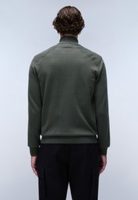 Grön tröja med hög krage och ribbade ärmslut, med texturerade axeldetaljer och en figurnära design. Bärs med svarta byxor.