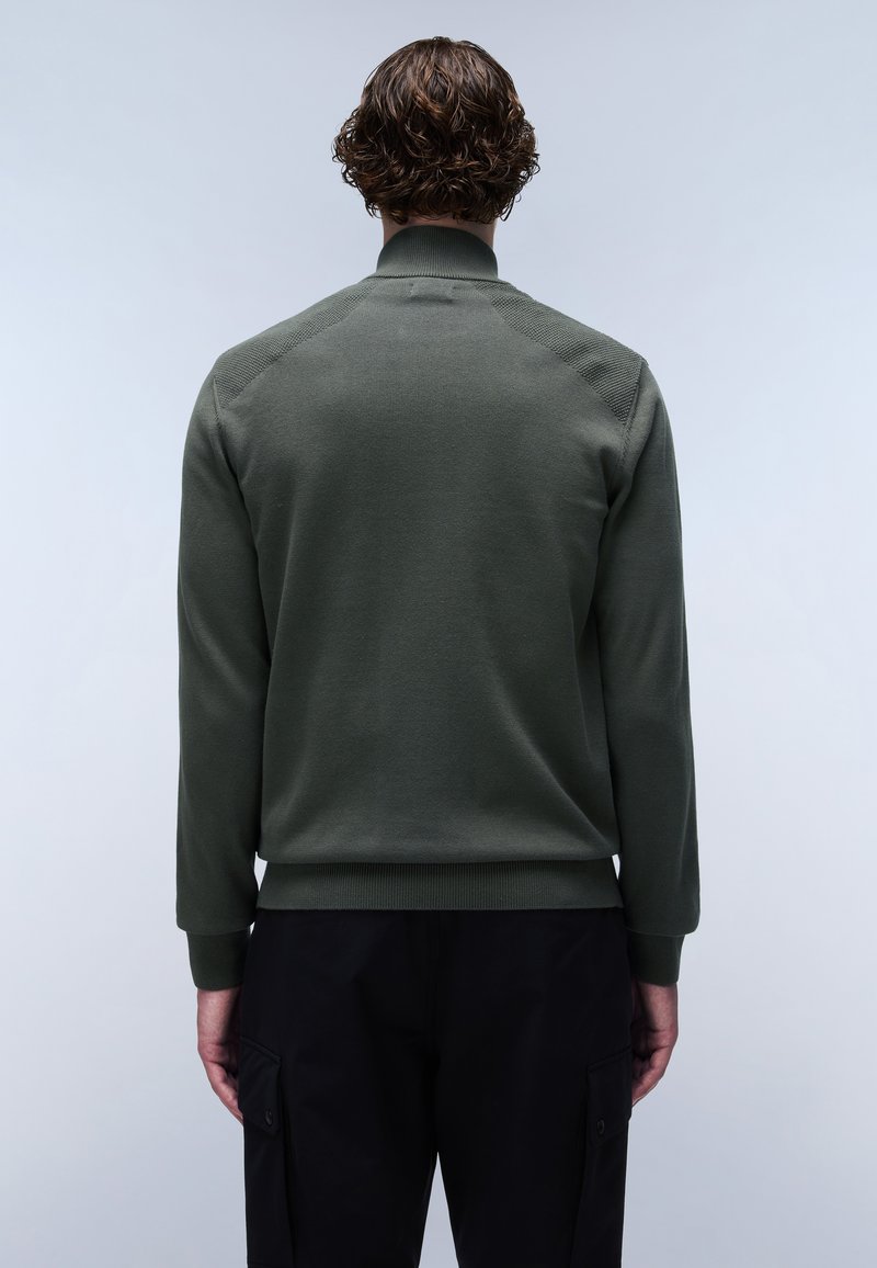 Pull vert avec un col haut et des poignets côtelés, présentant des accents texturés aux épaules et un design ajusté. Porté avec un pantalon noir.