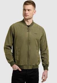 Homme portant une veste aviateur vert olive avec fermeture éclair et poches, debout avec une main dans la poche, regardant sur le côté.