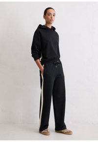 Zwarte oversized hoodie met trekkoord, gecombineerd met zwarte joggingbroek met crèmekleurige zijstrepen. Het model draagt lichtbruine sneakers.