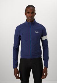 Marineblaue Radjacke mit einem Reißverschluss vorne, reflektierendem grauen Armreifen und Rapha-Logo. Hergestellt aus einem leichten, glatten Material.