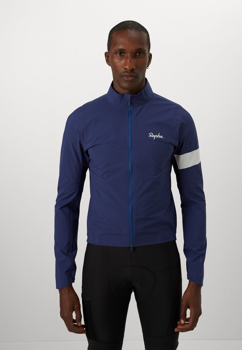 Marineblauwe fietsjacket met een ritssluiting aan de voorkant, reflecterende grijze armband en het Rapha-logo. Gemaakt van een lichtgewicht, glad materiaal.