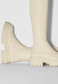 Botines beige hechos de material sintético suave, con una suela gruesa texturizada y paneles laterales elásticos para facilitar su uso.