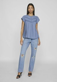 Blusa azzurra chiara a maniche corte con bordi arricciati e cuciture decorative, abbinata a jeans di denim chiaro strappati e tacchi aperti neri.