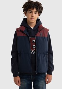 Navy zilā veste ar bordo augšējo daļu, satur kapuci un divas kabatas ar rāvējslēdzējiem. Izgatavota no mīksta, viegla auduma.