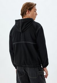 Zwarte hoodie met elastische manchetten en zoom, gekenmerkt door contrasterende witte stiksels. De achterkant toont een naaddetail over de schouders.