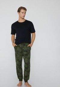 Man in een zwart shirt met korte mouwen en groene camouflage joggerbroeken, staat blootsvoets met de handen in de zakken, kijkt naar links.