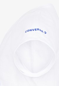 Camiseta de algodón blanca con escote redondo y logo en texto azul "CONVERSE" en la parte superior izquierda. Textura suave, diseño simple.