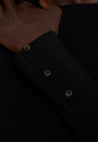 Poignet de pull côtelé noir avec trois boutons noirs, en matériau texturé qui ajoute une profondeur visuelle subtile.