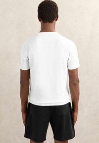Tricou alb cu mânecă scurtă și gât rotund, cu o textură netedă, purtat cu pantaloni scurți negri, evidențiind un design simplu și clasic.