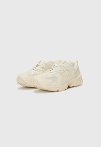 Lysebeige sportsko med mesh-paneler, polstret krage og teksturert gummisåle. Snørebånd og merking er subtilt integrert.