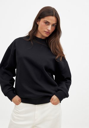 Sweatshirt noir avec un col rond, des épaules déchues et des poignets côtelés. Porté sur un pantalon blanc, avec une texture lisse.