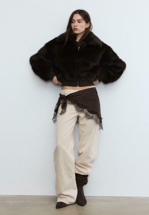 Bruine cropped jas van nep bont met ritssluiting, gecombineerd met crèmekleurige broek met wijde pijpen en een bruin wikkelstuk met kanten rand, wat textuur en contrast benadrukt.