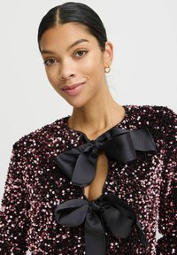 Sort sequin-top med rund halsudskæring, prydet med to store sorte satin-sløjfer foran og en tekstureret, glitrende overflade.