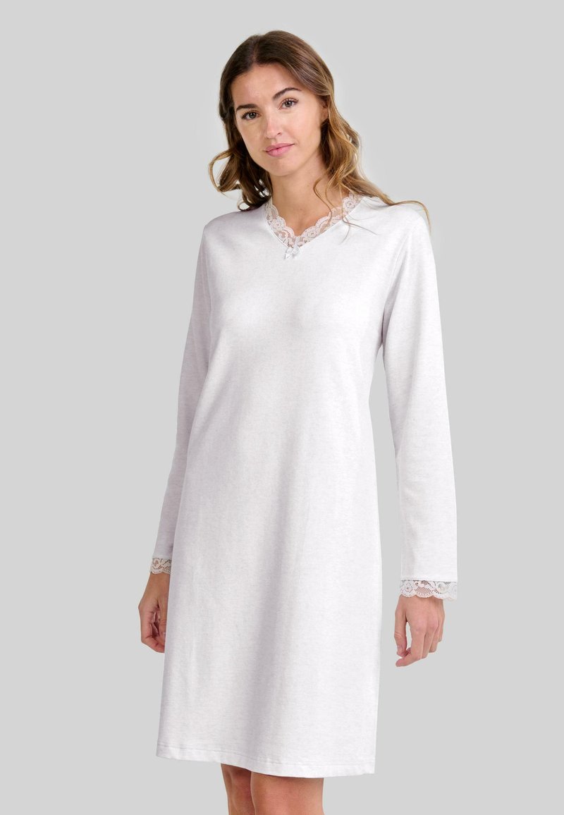 Mujer vestida con un camisón blanco de manga larga con encaje en el escote y los puños, de pie ante un fondo gris plano.
