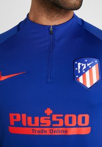 Sininen urheilullinen vetoketjupaita punaisilla yksityiskohdilla. Varustettu logopatchillä ja rohkealla tekstillä, jossa lukee "Plus500 Trade Online" alareunassa.