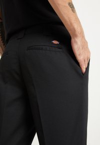 Černé kalhoty s hladkou texturou, obsahující jednu zadní kapsu a malou nášivku loga Dickies na pásku.