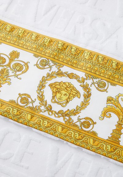 Serviette blanche avec une bordure décorative jaune et or audacieuse comportant des motifs complexes, y compris des feuilles de laurier et un motif de visage.