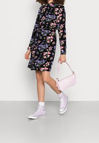 Blommig svart klänning med långa ärmar, bälte i midjan, rosa och lila blommor. I kombination med vita ankelsockor och lavendel Converse-sneakers, håller en rosa väska.