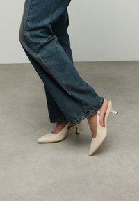 Beige leren slingback hakken met een puntige neus, voorzien van een dunne, gebogen hak en een open achterkant. Draagt met blauwe rechte jeans.