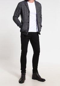 Persona che indossa un cardigan grigio scuro con bottoni, una maglietta bianca, pantaloni neri e stivali stringati marrone scuro, in piedi con le mani in tasca.