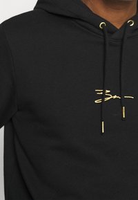 Sudadera negra de mezcla de algodón, con un pequeño logotipo bordado en dorado y aglets dorados en los cordones. Textura suave, diseño casual.