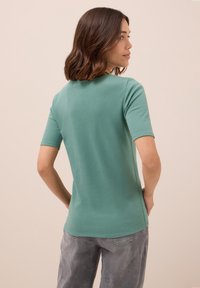 Eine Frau mit schulterlangem braunem Haar steht seitlich und trägt ein grünes kurzärmliges Oberteil und graue Jeans vor einem beigen Hintergrund.