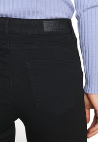 Jeans negros con un diseño estilizado, que presentan una etiqueta de cuero en la cintura, dos bolsillos traseros y un ajuste entallado. Se pueden ver detalles de costura mínimos.