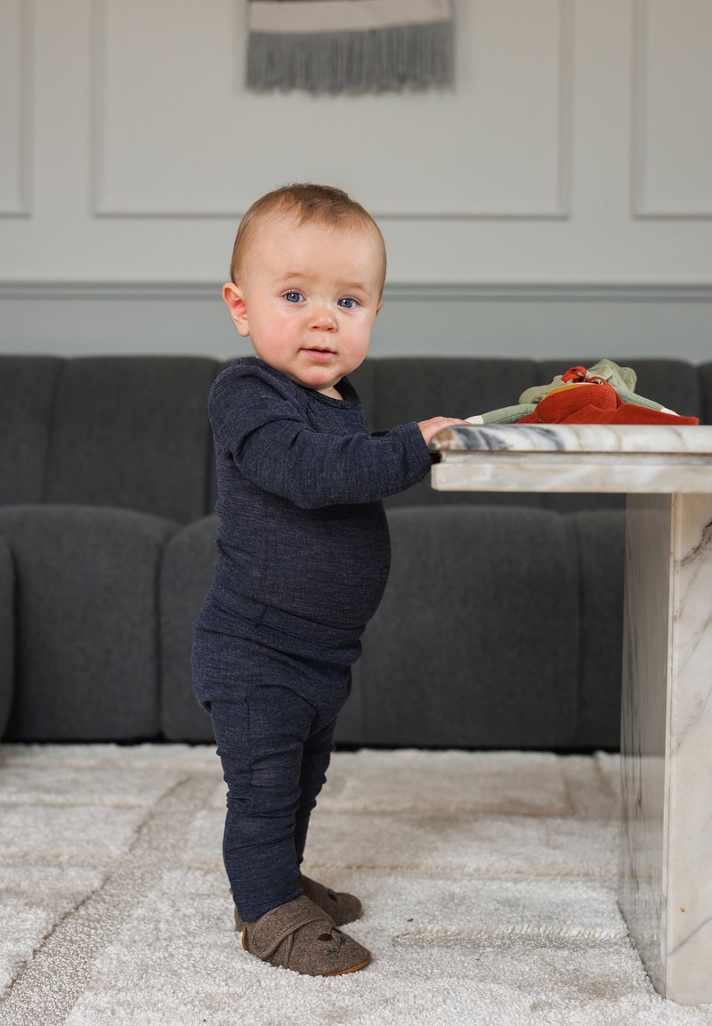 Sötétkék, hosszú ujjú rugdalózó álló kisgyermekön, bézs papucsokkal. Közvetlenül mellette márvány asztallap élénk, alatt puha szőnyeg. Háttérben szürke kanapé.