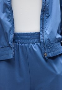 Μπλε ζακέτα με φερμουάρ, φορεμένη ανοιχτή πάνω από παντελόνι με ελαστική μέση στο ίδιο χρώμα και λευκό πουκάμισο με πέρφορέ λεπτομέρειες από κάτω.