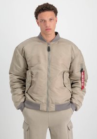 Chaqueta bomber beige con cierre frontal, puños acanalados, dos bolsillos laterales y una etiqueta en la manga. Tela suave y acolchada con un acabado satinado.