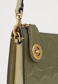 Gros plan d'un sac à main en cuir vert olive avec une quincaillerie dorée, un fermoir tournant et un motif circulaire embossé sur le côté.