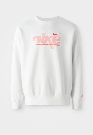 Bela majica iz bombažne mešanice. Ima velik rdeč "nike" logotip s potiskom pikic in spodaj besedilo "Mednarodna športna oblačila". Rdeči Nike swoosh na rokavu.