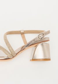 Sandale à talons en métal doré avec des sangles texturées et des accents brillants, dotée d'un talon bloc et d'une bride de cheville réglable.
