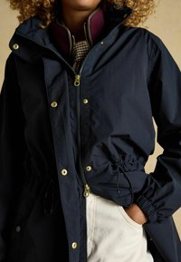 Jachetă impermeabilă bleumarin cu fermoar pe față, accesorii aurii, talie ajustabilă cu șnur și guler tip funnel căptușit cu material violet.