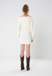 Vit stickad klänning med avtagna axlar, långa ärmar, figurnära design och mjuk textur, tillsammans med mörkbruna broderade klackstövlar.