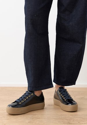 Personne portant des baskets compensées en cuir noir avec des semelles couleur cuir et un jean bleu foncé, debout sur un sol en bois.