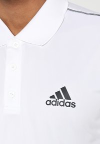 Bílá polo košile s hladkým povrchem, dvěma knoflíky u límečku a černým logem Adidas na levé straně. Černé stehy zvýrazňují límeček.