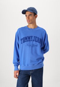 Modra kapuca z teksturiranim logotipom "TOMMY JEAN", okrogla izrezana ovratnik in rebrasti manšeti. V kombinaciji s temno modrimi jeans hlačami in usklajeno kapo.