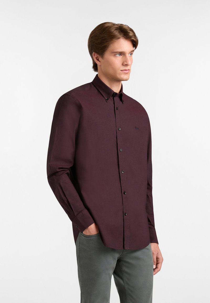 Giovane uomo con capelli castani che indossa una camicia a maniche lunghe viola scuro abbottonata e pantaloni grigi, con le mani in tasca, che guarda a destra.