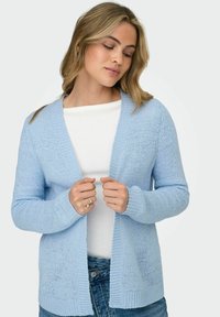 Woman wearing a light blue knitted cardigan over a white top and blue jeans, holding the cardigan edges with both hands.