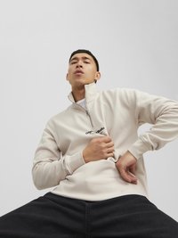Lys beige fleece pullover med lynlås-krave, med sort logo tekst. Modellen har mørke jeans på, siddende med hænderne på tøjet.