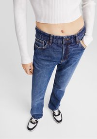 Blaue Jeans aus Denim mit geradem Beinschnitt, fünf Taschen und kontrastierenden Nähten. Kombiniert mit einem weißen gerippten Croptop und schwarzen Sneakers.