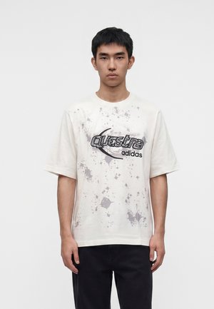 Jeune homme portant un T-shirt graphique blanc Adidas Questra avec des motifs éclaboussés noirs et gris, associé à un pantalon noir, sur fond blanc.