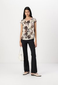 Blouse à manches courtes à motifs floraux en crème, associée à un pantalon noir évasé et des chaussures slip-on beiges. Elle présente une coupe décontractée et un col rond.