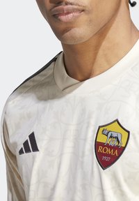 Camiseta de fútbol beige con un patrón sutil, que presenta un logo negro de Adidas en el hombro y un escudo del club Roma en el pecho.