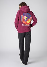 Bordeauxrode hoodie met een levendige bloemenprint op de achterkant, met diverse kleuren en patronen. Gecombineerd met donkergrijze broek en witte schoenen.