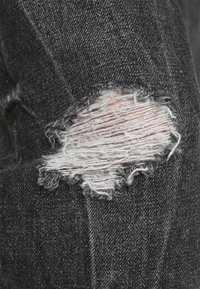 Tejido de mezclilla negro con un desgarro rectangular deshilachado que muestra hilos blancos y piel debajo del área dañada.