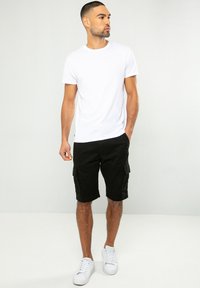 Weißes Kurzarm-T-Shirt kombiniert mit schwarzen Cargo-Shorts. Die Shorts verfügen über Taschen und sind knielang. Unten wurden weiße Sneakers getragen.