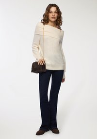 Maglione crema con scollo off-the-shoulder e colletto a costine, abbinato a jeans svasati scuri. Il modello porta una borsa a tracolla in ecopelliccia marrone.